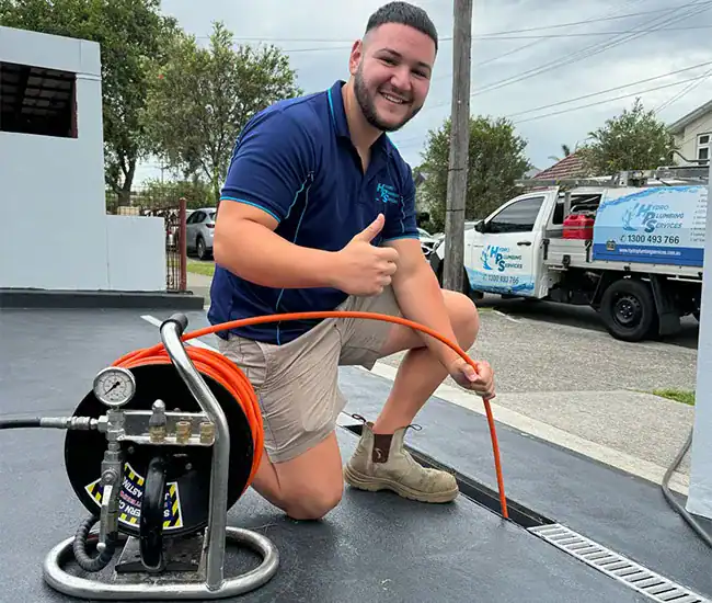A smiling worker in a blue shirt and beige shorts kneels on the ground and gives a thumbs up. He holds an orange hose that is connected to a piece of equipment on the left. A company van, showcasing Plumbers Sydney, is parked in the background on a suburban street.