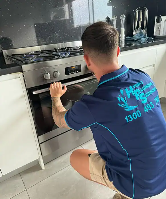 A person wearing a "HPR Plumbing Services" polo shirt is kneeling and checking an oven in a modern kitchen. With short hair and visible tattoos on their arm, they expertly handle appliance installations. The kitchen features white cabinets, a gas stove, and various items on the counter.