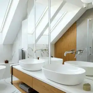 A modern bathroom in Sydney with a sloped ceiling features two large, circular white sinks on a wooden vanity with open shelves. Two large mirrors hang above the sinks, complemented by a marble backsplash and skylight windows letting in natural light. A bathtub is visible in the background, showcasing recent renovations.