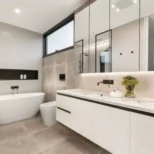 Modern bathroom renovation in Sydney featuring a white freestanding bathtub, a white toilet, and a double-sink vanity with large mirrors. The walls boast neutral tiles while large windows flood the space with natural light. A vase of flowers on the vanity adds a touch of color.
