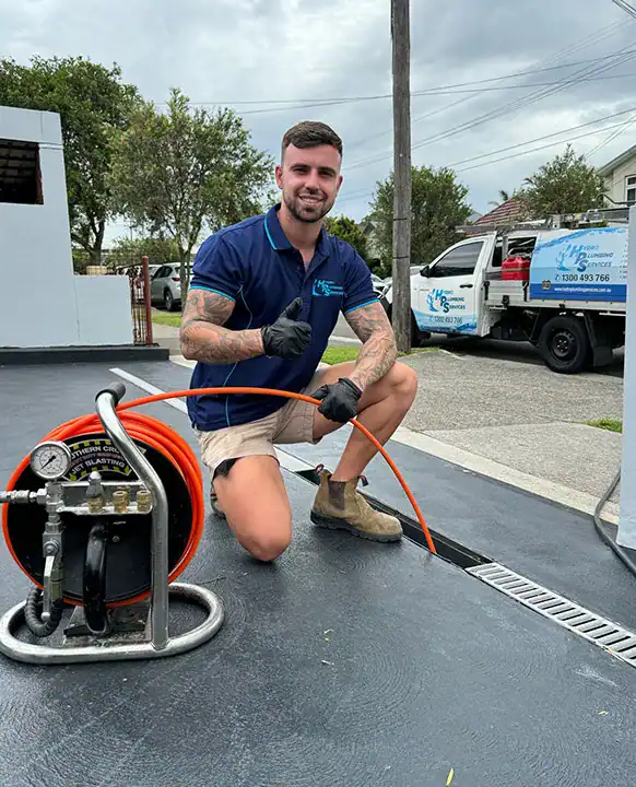 A man kneels on a paved surface, smiling and giving a thumbs-up. He wears a blue polo shirt, khaki shorts, gloves, and work boots. He's holding an orange hose connected to a machine used for clearing blocked drains in Sydney. A utility truck with a business logo is in the background. Trees and cloudy sky are visible.