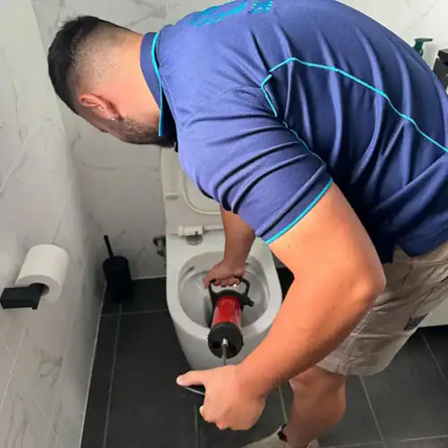 A person in a navy blue shirt and khaki shorts uses a red plunger to unclog a toilet in a modern bathroom with white tiled walls and dark floor tiles. The bathroom, reminiscent of those seen in blocked drains Sydney ads, also features a toilet paper holder and a toilet brush nearby.