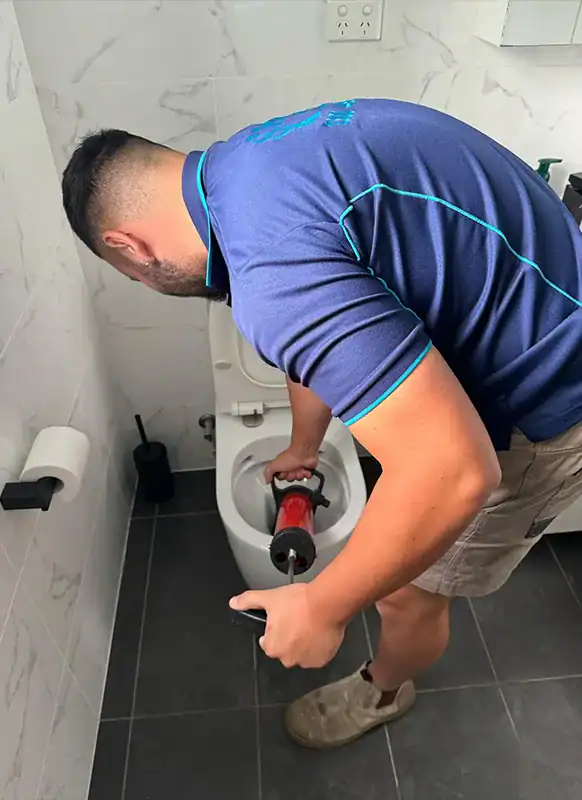 A person in a navy blue shirt and khaki shorts uses a red plunger to unclog a toilet in a modern bathroom with white tiled walls and dark floor tiles. The bathroom, reminiscent of those seen in blocked drains Sydney ads, also features a toilet paper holder and a toilet brush nearby.