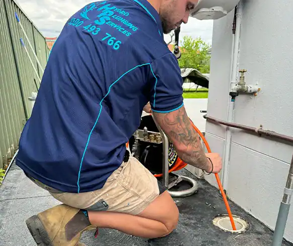 A plumber in a blue and black uniform with visible tattoos on his arm is kneeling and inserting an orange hose into a drainage pipe on the ground. Various tools and equipment are around him, highlighting his expertise in All General Plumbing. A fenced area and greenery are visible in the background.