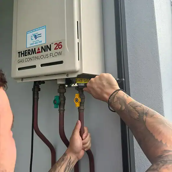 A person with tattooed arms is adjusting or inspecting the connections on a "Thermann 26 Gas Continuous Flow" hot water system mounted on an exterior wall. Various colored valves and gas lines are visible connected to the unit.