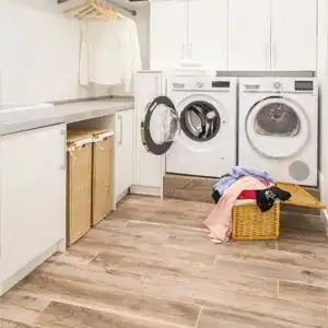 A clean laundry room with wooden flooring, fresh from a recent renovation in Sydney, features a washing machine and dryer side by side. The washer door is open with towels inside. A basket of laundry sits on the floor, and cabinets and shelves hold additional items and hangers.