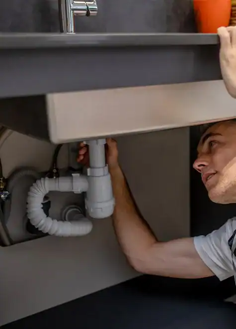 A person inspects the plumbing under a kitchen sink, reaching up to touch the pipes, which include a white P-trap and various connectors. Concerned about blocked drains, they examine each part closely. The countertop and faucet are clearly visible above in the bright lighting that illuminates the details of the plumbing.