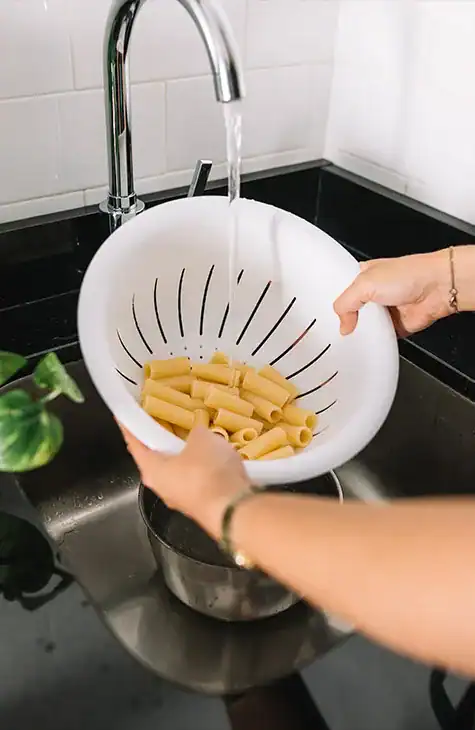 A person rinses uncooked rigatoni pasta in a white colander under running water from a faucet in a kitchen sink, taking care to avoid blocked drains. The kitchen has white tiled walls and a black countertop. A small green plant is partially visible to the left of the sink.