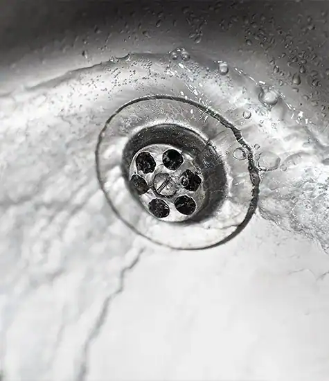 Close-up image of a metal sink drain with water flowing. Water droplets are visible around the drain, creating a dynamic, swirling effect often seen in blocked drains. The photo captures the texture of the stainless steel and the motion of the water.