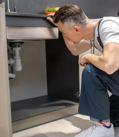 A man wearing a white shirt and grey overalls kneels and closely inspects the plumbing under a kitchen sink. The cupboard doors below the sink are open, revealing the pipes, possibly assessing for blocked drains.