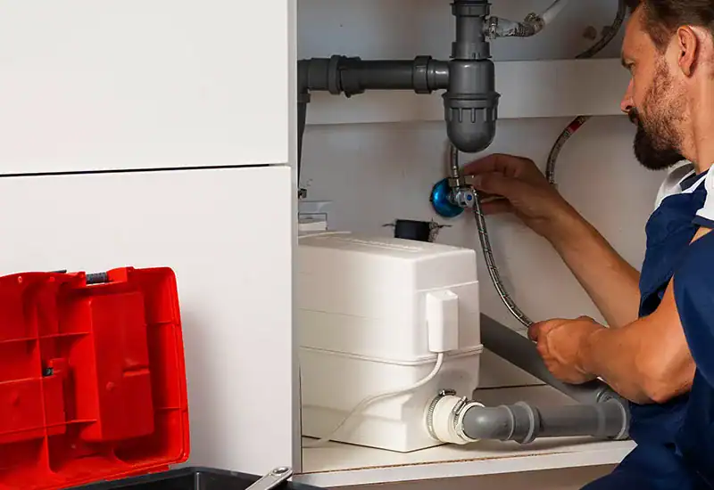 A plumber in overalls works under a kitchen sink, adjusting pipes to clear blocked drains, using tools from an open red toolbox placed nearby. The kitchen cabinets are white, and the plumber appears focused on the repair work.