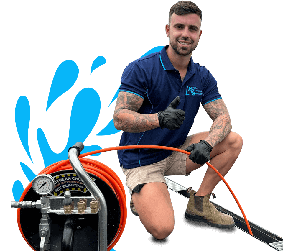 A man in a blue polo shirt and tan shorts kneels while giving a thumbs up. He is holding an orange hose connected to a pressure gauge and control panel, suggesting a cleaning or blasting service. With tattoos on his arms, gloves, and work boots, he looks every bit the professional plumber Sydney trusts.