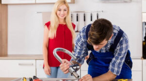A Sydney plumber wearing blue overalls is fixing a kitchen faucet while a woman in a red blouse watches in the background. Various tools are scattered on the kitchen counter next to them.