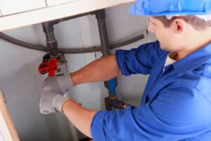 A plumber near me, wearing a blue helmet and blue uniform, works diligently with plumbing pipes under a sink, using a red wrench. The person is also wearing protective gloves while adjusting or fixing the pipes.