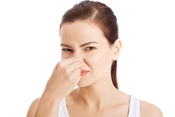 A woman with a displeased expression pinches her nose with her fingers, seemingly reacting to an unpleasant smell. She has dark hair pulled back and is wearing a white sleeveless top. The plain white background emphasizes the likely issue of faulty bathroom plumbing causing the odor.
