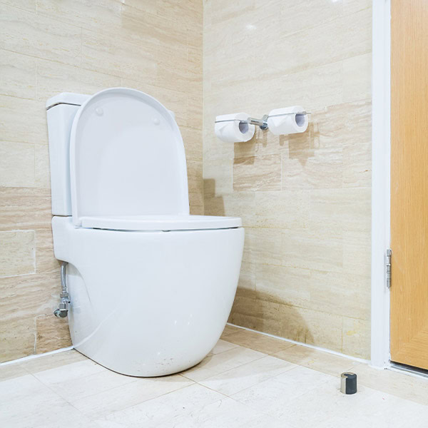A modern, porcelain toilet with the seat lid open is positioned against a beige tiled wall. Two empty toilet paper holders are mounted on the wall to the right. If you're facing issues, consider contacting Toilet Repairs Sydney for prompt solutions. The bathroom floor is also covered with beige tiles.