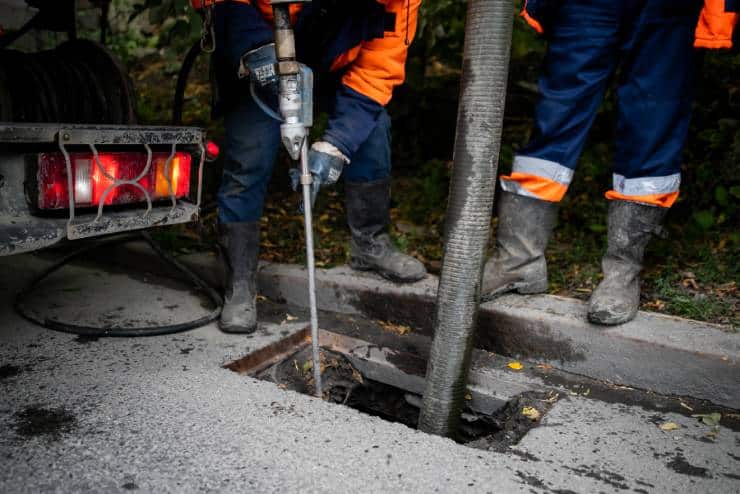 Two workers wearing orange safety jackets and blue pants operate industrial equipment. One man is holding a high-pressure hose, performing jet cleaning, while the other manages a large suction hose inserted into an open sewer hole in the street. A service truck is parked nearby.