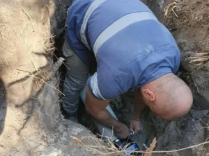 A person, likely a plumber, wearing a blue shirt and khaki pants is seen from behind, crouching and working in a dug-out hole. The individual appears to be fixing a pipe or some other equipment in the ground. The surrounding area has exposed soil and roots, typical of a day&rsquo;s work in Sydney.