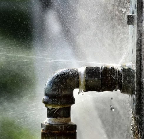 Close-up of a metal pipe joint spraying water from multiple small leaks. The pipe is weathered and the escaping water is caught mid-action, creating a misty, damp scene. This image highlights issues similar to common running toilet problems. The background is blurred, focusing attention on the leaking pipe.