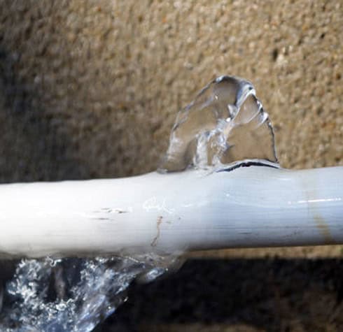 A white pipe with a visible crack has water spraying out of it forcefully. The background is a textured surface, possibly a wall. The image showcases common running toilet issues due to a damaged pipe causing significant water leakage.