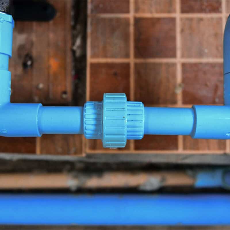Close-up of a section of blue PVC plumbing pipes connected by a plastic coupling joint, potentially part of a jet cleaning plumbing system. The pipes form a U-shape and are installed against a backdrop of brown and beige ceramic tiles with some visible wear. Additional blue pipes are seen in the background.