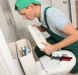 A plumber in green overalls and a cap, with tools nearby, is lifting the lid off a toilet tank, focused on fixing or inspecting the internal components to address common running toilet issues in a bathroom.