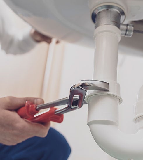 A person uses red pliers to tighten a white plastic pipe under a sink. The scene, reminiscent of reliable appliance installation services, focuses on the hands and the plumbing work, with the background slightly blurred.
