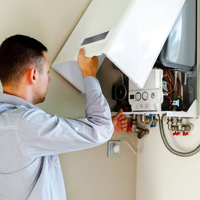 A man in a light gray shirt is servicing a wall-mounted boiler. He is removing the front panel to access the internal components for hot water system inspection services. Various pipes and gauges are visible, indicating the boiler's plumbing and control systems. The setting appears to be indoors.