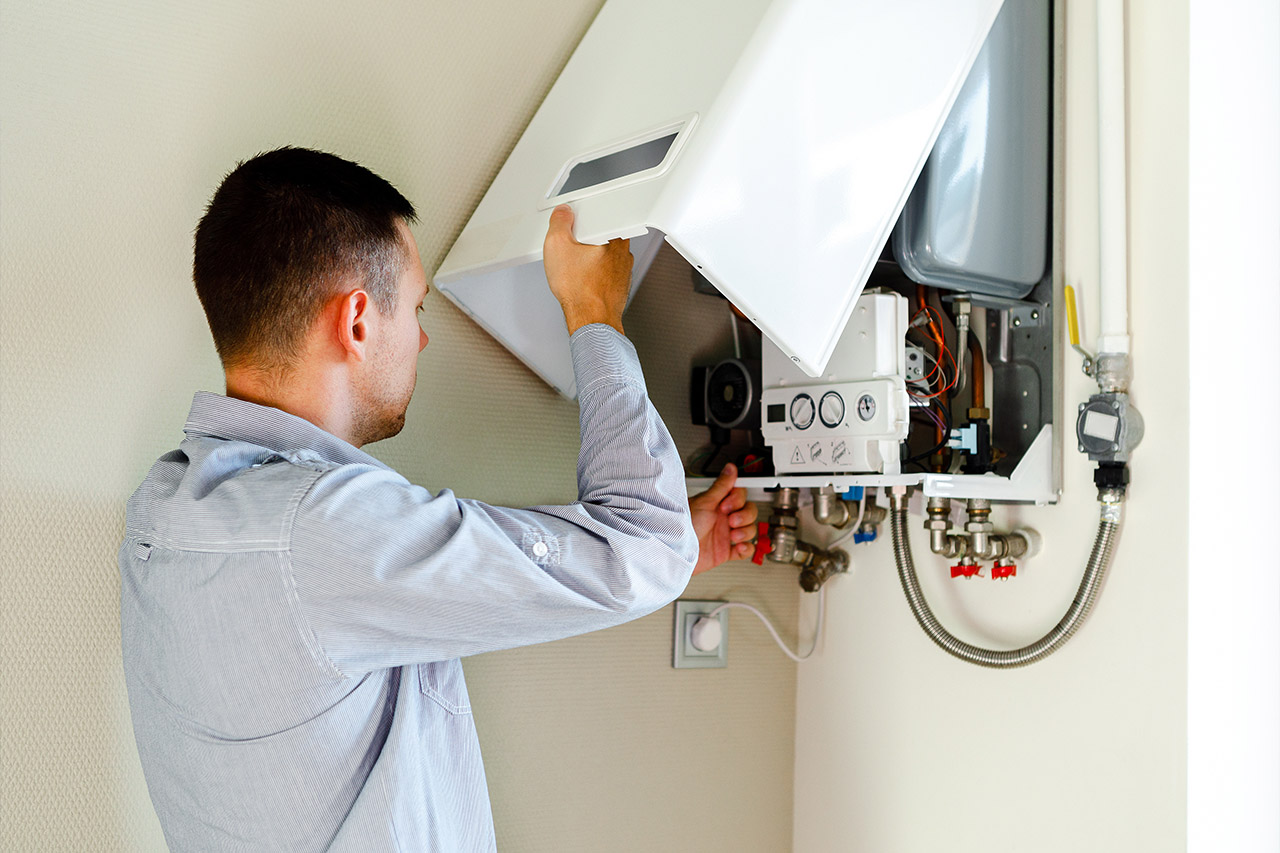 A man in a light gray shirt is servicing a wall-mounted boiler. He is removing the front panel to access the internal components for hot water system inspection services. Various pipes and gauges are visible, indicating the boiler's plumbing and control systems. The setting appears to be indoors.