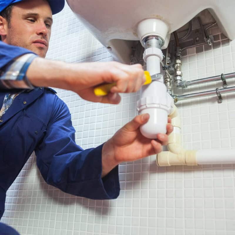 A plumber in a blue uniform and cap uses a wrench to tighten the pipes under a sink in a tiled bathroom, ensuring top-notch plumbing services.