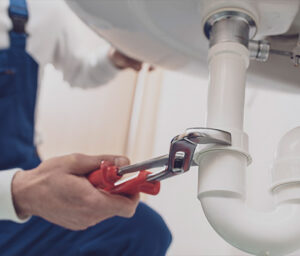 A plumber in blue overalls uses a red pipe wrench to tighten white PVC pipes under a sink. Only the plumber's hands and part of the upper body are visible, with focus on the meticulous plumbing inspection services being provided.
