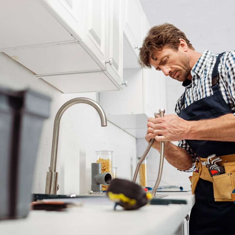 Need a reputable plumber? A man in a checkered shirt and apron diligently works on the plumbing under a kitchen sink, holding a flexible pipe. A toolbox sits in the foreground, with kitchen cabinets above him. A tool belt brimming with various tools is snug around his waist.