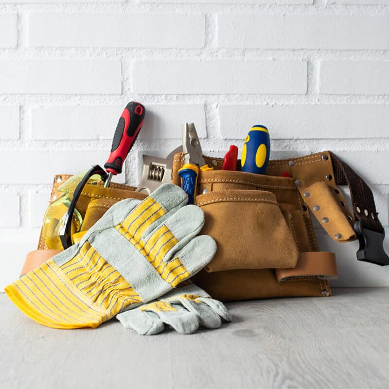 A leather tool belt is resting on a light wood floor with a white brick wall backdrop. The belt is filled with various tools, including pliers, screwdrivers, and a measuring tape—everything you might need to address common household issues like improving water pressure in your home. A pair of gray and yellow work gloves are placed in front of the belt.