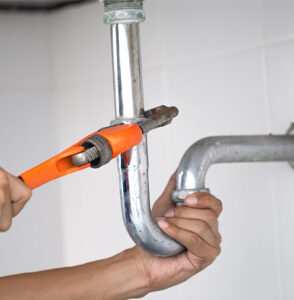 A person uses an orange-handled wrench to tighten or loosen the connection on a shiny metal pipe under a sink, possibly addressing concerns like "How Can I Improve Water Pressure in My Home." The person's hands grip both the pipe and wrench, clamped around the pipe's curved section. The surrounding area is tiled in white.