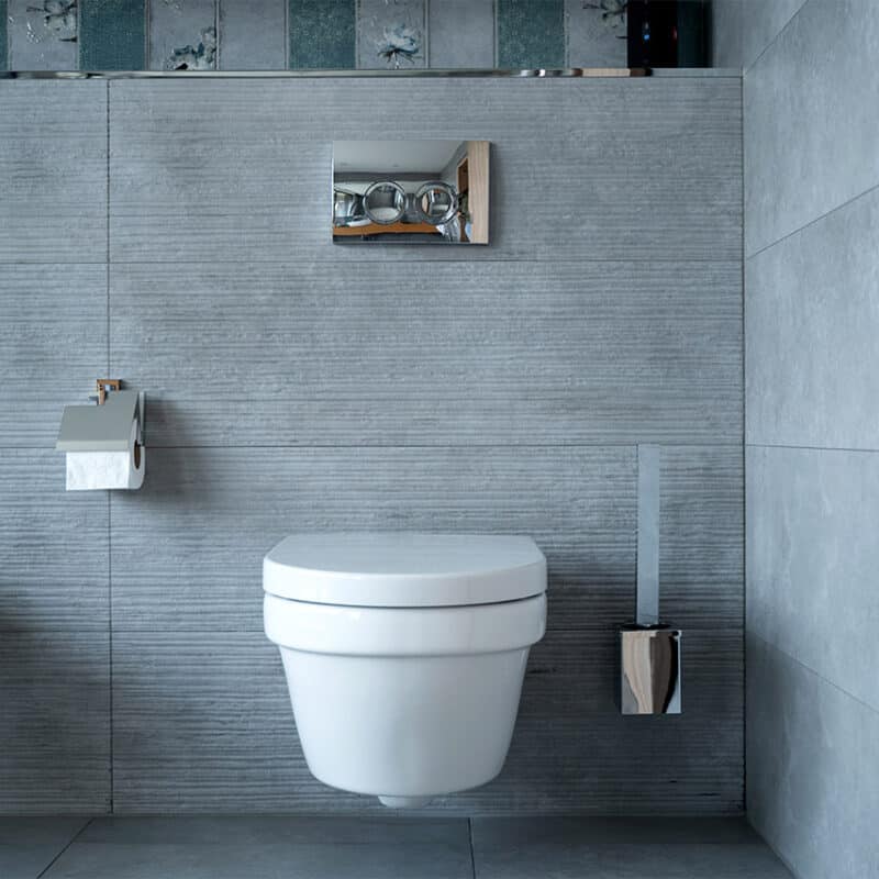 A modern bathroom with light gray textured walls and flooring features a wall-mounted toilet adorned with a chrome flush button. Next to it, there's a chrome toilet paper holder and a matching brush holder. If you find yourself wondering, "Can I repair a broken toilet or should I replace it?" this setup provides sturdy fixtures worth keeping.