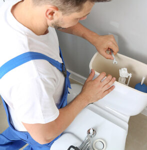 A plumber wearing blue overalls uses a wrench to repair the inside of a toilet tank, contemplating the age-old question, "Can I Repair a Broken Toilet or Should I Replace It?" He is standing beside the toilet, focusing on adjusting the internal components. The tank lid is removed, and a plunger can be seen in the background.