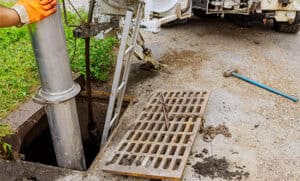 A worker in orange gloves is conducting plumbing maintenance, using equipment to clean a sewer drain. The metal grate from the drain is lifted and placed to the side, attached with a chain. There is a ladder leading down into the sewer, and a blue tool is on the ground nearby.