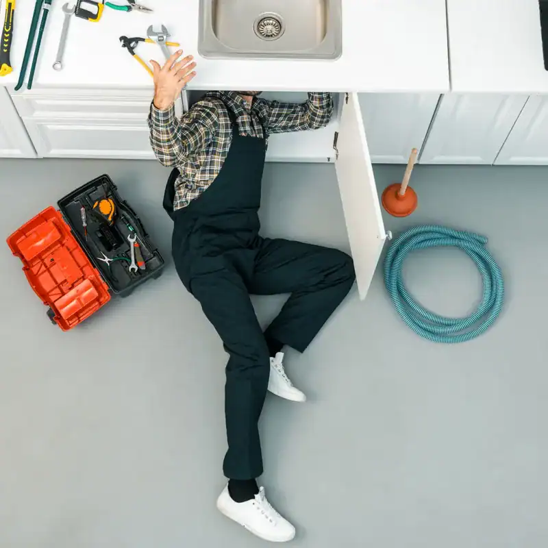 A plumber in Sydney dressed in overalls and a plaid shirt is lying on the floor, working under a kitchen sink. A red toolbox with tools, a plunger, and a coiled hose are nearby on the kitchen floor. The surrounding countertops include a sink and a stove.