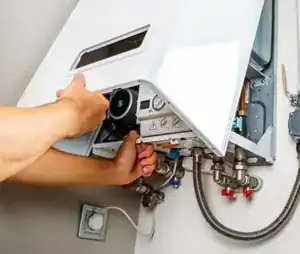 A person is performing maintenance on a wall-mounted gas boiler. The front panel of the boiler is open, revealing various pipes, knobs, and internal components. The person's hands are adjusting parts of the system, showcasing the expert skills of professional plumbers in Sydney.
