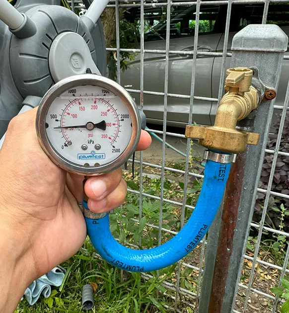 A hand holding a pressure gauge connected to a blue hose and a brass valve attached to a metal pipe, with the gauge displaying readings in PSI from 0 to 200. The setup, which looks like it's part of an inspection by a Sydney emergency plumber, appears to be near a wire fence and a parked car.