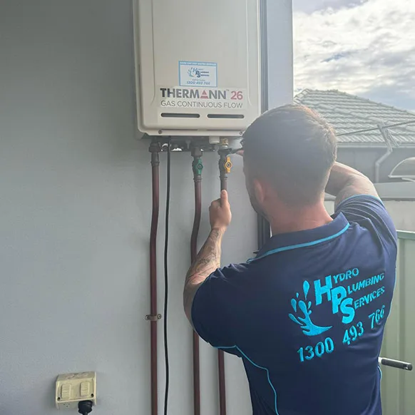 A plumber wearing a blue shirt with "Hydro Plumbing Services" on the back is adjusting a pipe connected to a Thermann 26 model outdoor gas continuous flow hot water system mounted on a wall.