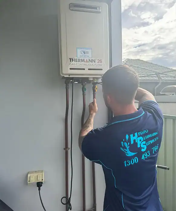 A plumber in a blue uniform is working on a wall-mounted gas continuous flow water heater. As one of the skilled gas fitters, he uses a wrench to adjust a valve on the pipes connected to the unit. The back of his shirt reads "Hydro Plumbing Services" with a phone number below.