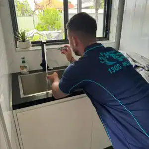 A plumber in a blue uniform kneels at a kitchen sink, working on the faucet. The sink area is clean, with a small potted plant on the countertop and a bottle of liquid soap nearby. Sunlight filters in through the window above the sink, hinting at his expertise beyond just toilet repairs Sydney loves.