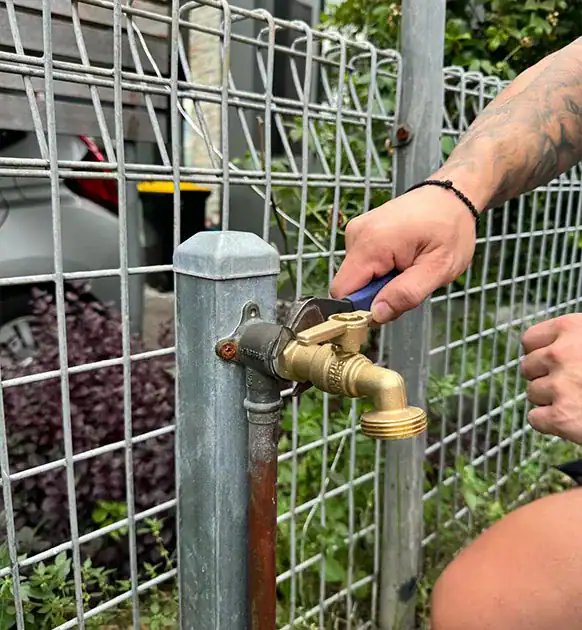 A person with a tattooed arm uses a wrench to tighten a brass outdoor faucet attached to a metal post. The scene, set against a wire fence with greenery in the background and parts of a vehicle visible behind it, suggests they're well-versed in various plumbing tasks like Toilet Repairs Sydney.