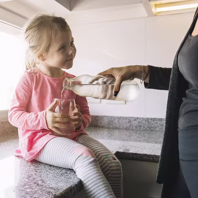 little girl on kitchen counter holding glass which is being poured water into by a woman wearing black dress.