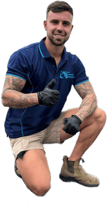 A man with a beard, wearing a blue shirt, light shorts, and sunglasses, sits on a wooden bench outdoors. His left arm rests on his knee while his right hand touches his chin. He has a relaxed expression, looking like he&rsquo;s waiting for a call from the Sydney emergency plumber, against a backdrop of greenery.